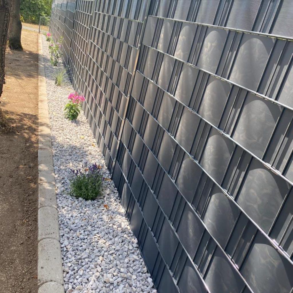 Anthrazitfarbene Sichtschutzstreifen im Streiflicht, davor ein Kiesstreifen mit Stauden